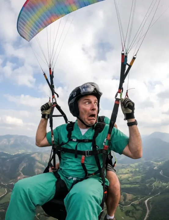 Dentist in green scrubs paragliding over a forested valley, mid-panic